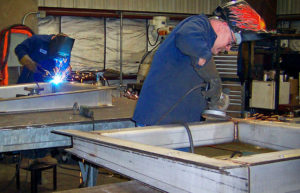 A man welding in an industrial setting with other workers.
